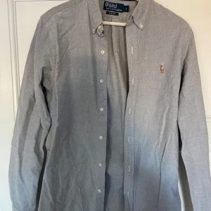 Grå skjorta från Polo Ralph Lauren - Säljer en stilren grå skjorta från Polo Ralph Lauren i slim fit. Skjortan har långa ärmar och knappar framtill. Perfekt för både vardag och festliga tillfällen. Den är i mycket bra skick och har en liten broderad logga på bröstet.