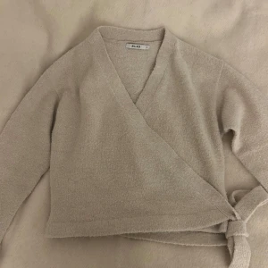 Beige omlottkofta från NA-KD - Mysig beige omlottkofta från NA-KD i storlek S. Perfekt för höst och vår med sin mjuka och bekväma passform. Koftan har långa ärmar och knytning i sidan för en stilren look. Passar både till vardags och lite finare tillfällen.