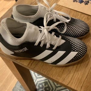 Adidas Predator fotbollsskor - Säljer ett par Adidas Predator fotbollsskor i svart och vitt. Skorna har en snygg design med vita ränder och ett mönster på ovansidan. De är i mycket bra skick och perfekta för inomhusfotboll. Tillverkade i syntetmaterial med snörning för bra passform.