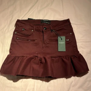 Vinröd kjol från Scotch & Soda - Snygg vinröd kjol från Scotch & Soda i jeansmaterial. Kjolen har en volangdetalj längst ner och flera fickor med dragkedjor fram. Perfekt för höst och vår, och kan stylas både upp och ner beroende på tillfälle. Helt ny med taggar kvar! Köpt för 1200.