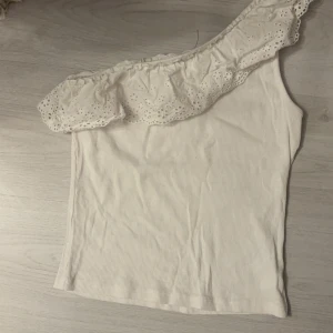 Vit oneshoulder topp från Kappahl - Säljer en superfin vit oneshoulder topp från Kappahl. Den har en söt volangdetalj längs axeln och är ribbad för en skön passform. Perfekt för sommarens alla fester eller bara en solig dag ute. 🕶️