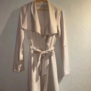Beige trenchcoat med knytband - Säljer en elegant beige trenchcoat med knytband i midjan. Jackan har långa ärmar och en stilren design med bred krage. Perfekt för vår och höst när du vill ha en chic look. Passar både till vardags och festliga tillfällen.