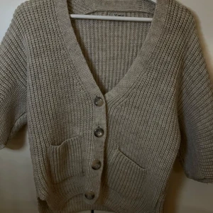 Beige stickad kofta med knappar - Mysig beige stickad kofta med stora knappar framtill. Den har en djup v-ringning och praktiska fickor. Perfekt för kyliga dagar när du vill vara både bekväm och stilren. Passar bra till både jeans och kjol!