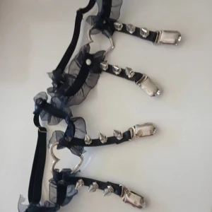 Svart bälte med spets och nitar - Säljer ett unikt svart strumpklämmor med coola detaljer! Det har spetsiga nitar och en hjärtformad metallring i mitten. är dekorerat med svart volangspets som ger en extra edgy look. Perfekt för att ge din outfit en punkig touch!