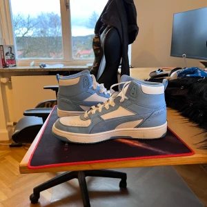 Blå och vita sneakers från Puma - Säljer ett par snygga blå och vita sneakers från Puma i mycket bra skick. De har en klassisk hög design med snörning och Puma-loggan på plösen. Perfekta för både vardag och träning. Skorna är bekväma och stilrena, ett måste för sneakerälskare! 800 kr nya. De är använda 1 gång.