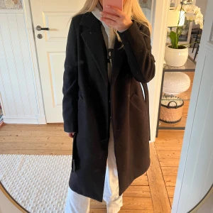 Svart kappa med bälte - Säljer en elegant svart kappa i mycket bra skick. Den har långa ärmar och ett stilrent bälte i midjan för en snygg siluett. Perfekt för höst och vår, och funkar lika bra till vardags som till fest. Kappan är i ett klassiskt snitt och passar till det mesta i garderoben.