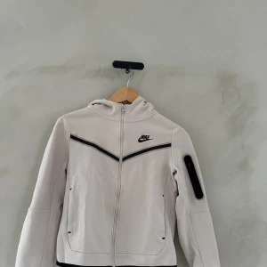 Vit vindjacka från Nike - Säljer en snygg vit vindjacka från Nike med dragkedja och huva. Jackan har långa ärmar och en cool svart detalj på ärmen. Perfekt för vår och höst när det blåser lite extra. Passar både till träning och vardagsoutfits. 🏃‍♂️