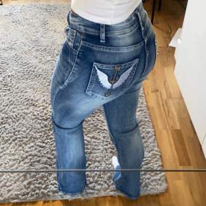 Säljer ett par snygga blå jeans i mycket bra skick. De har en normal passform och är lågmidjade. På bakfickorna finns coola broderade vingar som ger en unik touch. Perfekta för både vardag och fest!