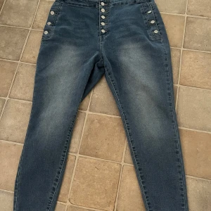 Blå jeans från Shein Curve - Säljer ett par snygga blå jeans från Shein Curve i storlek 1XL. De är högmidjade med en rad knappar framtill och dekorativa knappar vid fickorna. Jeansen har en klassisk femficksdesign och är perfekta för både vardag och fest. Materialet är en blandning av polyester och elastan för en bekväm passform.
