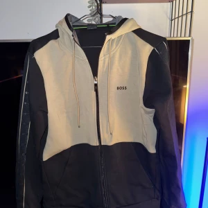 Svart och beige hoodie från BOSS - Säljer en snygg svart och beige hoodie från BOSS. Den har en dragkedja framtill och en justerbar huva med snören. Perfekt för höst och vår, med en stilren design som passar både till vardags och lite mer avslappnade tillfällen. Stor logga på bröstet ger en extra touch av stil.pris går att diskutera 