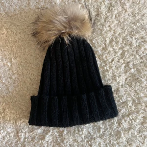 Mössa med pälsboll - Svart ribbad mössa med avtagbar pälsboll upptill.