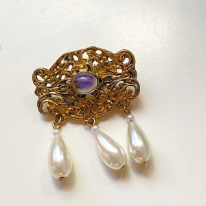 Vintage brosch med pärlor  - Säljer en superfin vintage brosch i guld med en  sten i mitten och tre hängande pärlor. Perfekt för att ge en elegant touch till din outfit. Broschen har ett intrikat mönster och är i bra skick. Den är cirka 5 cm bred. Passar perfekt till fest eller speciella tillfällen!