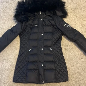 Svart dunjacka från Rockandblue - Säljer en snygg svart dunjacka från Rockandblue i storlek 36 och en likadan i 38. Jackan har en quiltad design med en fluffig huva. Den har dragkedja och knappar framtill samt flera praktiska fickor. Perfekt för kalla vinterdagar! ❄️