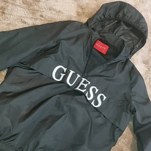 Svart vindjacka från Guess - Säljer en snygg svart vindjacka från Guess med stor logga på bröstet. Jackan har en huva och är perfekt för blåsiga dagar. Den är använd 6 gånger. Passar både till vardags och lättare utomhusaktiviteter.