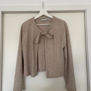 Beige kofta - Beige kofta ifrån Laget 157 i storlek M. Koftan passar till alla tillfällen och är perfekt nu när de börjar bli kallare ute.Koftan är grå och har två band fram till som man kan knyta till en rosett som på bilden eller bara ha hängandes som dem är. 