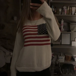 Vit stickad tröja med amerikansk flagga - Säljer en vit stickad tröja med en stor amerikansk flagga på framsidan. Tröjan har långa ärmar och en bekväm passform, perfekt för kyliga dagar. Den är både stilren och iögonfallande, idealisk för en casual look.