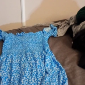 Blå blommig klänning - Söt blå klänning med blommigt mönster. Perfekt för sommardagar eller en avslappnad vårutflykt. Den har korta ärmar och en bekväm passform. Materialet känns lätt och luftigt, vilket gör den idealisk för varmare väder.