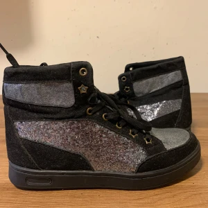 Svarta sneakers med glittriga detaljer - Säljer ett par coola svarta höga sneakers med glittriga silverdetaljer. Skorna har snörning och små stjärnor vid snörhålen som ger en unik touch. Perfekta för att lysa upp vilken outfit som helst! 🖤✨använd 1-2 gånger max