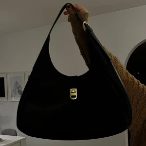 Svart axelväska med gulddetaljer - Snygg svart axelväska i skinn med elegant guldfärgad låsdetalj framtill. Perfekt för både vardag och fest. Väskan har en enkel och stilren design med en bekväm axelrem.