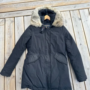 Svart parkajacka från Woolrich - Säljer en svart parkajacka från Woolrich i mycket bra skick. Jackan har en mysig pälsfodrad huva och långa ärmar. Perfekt för kalla vinterdagar. Den har praktiska fickor och stängs med dragkedja och knappar. Supervarm och stilren!