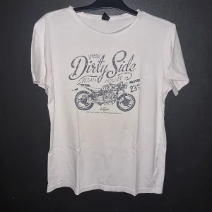 Vit t-shirt med motorcykeltryck - Säljer en vit t-shirt med coolt motorcykeltryck och texten 'Dirty Side'. Perfekt för en avslappnad stil. T-shirten är i bomull och har korta ärmar. Den har en liten reva på baksidan och missfärgad i armhålorna. 