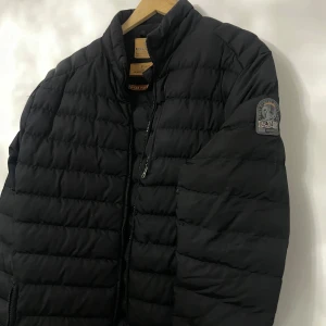 Svart dunjacka från Parajumpers - Säljer en snygg svart dunjacka från Parajumpers i mycket bra skick. Jackan är quiltad och har en dragkedja framtill. Perfekt för höst och vinter, med en normal passform och långärmade ärmar. Märkeslogga på ärmen ger en extra touch av stil. Perfekt för att hålla sig varm och trendig!