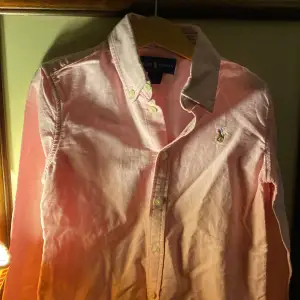 Säljer en snygg rosa skjorta från Ralph Lauren. Skjortan har långa ärmar och klassiska knappar framtill. Den är gjord i bomull och har en broderad logga på bröstet. Obs det är en barntröja som är rekommenderad för 8 åring. 