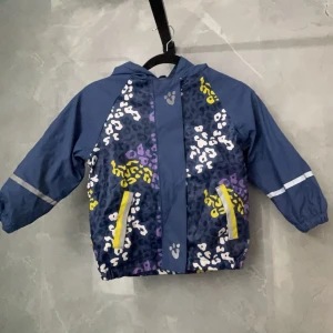 Blå mönstrad vindjacka med huva - Säljer en blå regnjacka  med huva och färgglatt mönster i vitt, gult och lila. Jackan har dragkedja framtill och två gula fickor. Perfekt för blåsiga dagar!