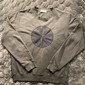 Grå sweatshirt från Stone Island - Säljer en stilren grå sweatshirt från Stone Island med ett broderat cirkelmönster på framsidan. Tröjan har långa ärmar och ribbade muddar vid ärmslut och nederkant och är av storlek L. Perfekt för en avslappnad stil.                                                                                                            Själv så har jag storlek M, Och den passar perfekt på mig. På bild 4 kan man se ett litet hål på armen. Hålet syns knappt när man har på den.
