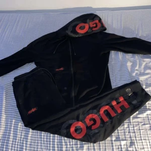 Svart hoodie och byxor från Hugo - Säljer en svart hoodie och matchande byxor från Hugo. Hoodien har en dragkedja och stor logga i rött på huvan. Byxorna har också en stor röd logga längs benet. Perfekt för en avslappnad stil.