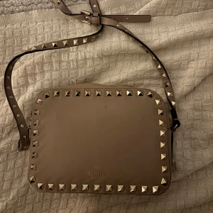 Beige axelväska från Valentino - Snygg beige axelväska från Valentino med nitar runt kanterna. Väskan har en justerbar axelrem och en dragkedja upptill. Perfekt för att ge din outfit en edgy touch. Väskan har inga defekter förutom att den är lite smutsig på insidan av botten men det syns inte om man har grejer i väskan. Pris går att diskutera 