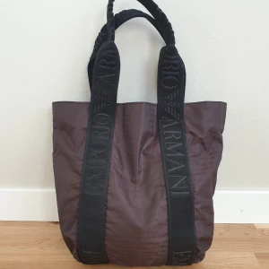 Brun shoppingväska från Emporio Armani - Snygg brun shoppingväska från Armani med svarta handtag där märkesnamnet är broderat. Väskan har en rymlig insida och är perfekt för att bära med sig allt man behöver. Tillverkad i ett slitstarkt material som ger en stilren look. 27×33×20cm