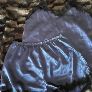 Blå satinlinne och shorts med spetsdetaljer - Säljer ett elegant blått satinlinne och shorts med svarta spetsdetaljer längs halsringningen. Linnet har en glansig finish och tunna axelband, perfekt för en lyxig känsla.