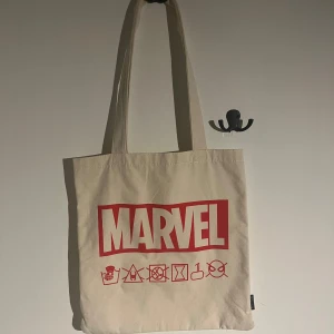 Beige tygväska med Marvel-tryck - Säljer en beige tygväska med rött Marvel-tryck och ikoner av olika superhjältar. Väskan har långa handtag och är perfekt för att bära med sig allt man behöver. Enkel och stilren design för alla Marvel-fans. Aldrig använd 💕