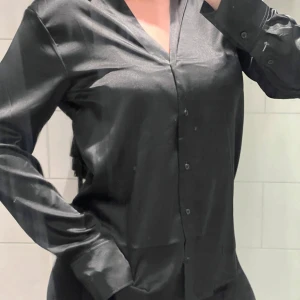 Svart sidenblus - Elegant svart blus i siden med långa ärmar och knappar framtill. Perfekt för en stilren look.