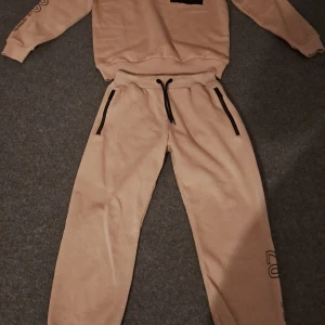 Beige Tracksuit with Black Details - Säljer en beige sweatshirt och matchande byxor från Identify. Tröjan har en svart ficka med dragkedja på bröstet och byxorna har svarta detaljer vid fickorna. Perfekt för en avslappnad stil.