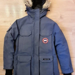 Canada Goose Expedition dunjacka - Canada Goose Expedition Parka i färgen Spirit, en vacker blå nyans. Avtagbar pälskrage, ståltråd i huvan, flera praktiska fickor och en justerbar midja. Jackan har några år på nacken men är välbevarad. Den har de klassiska tecknen på användning dvs slitage längst ner på mudden (hål i den ena mudden, se bild!), samt lite slitage, mindre hål längst med sömmarna på ärmarna (sista bilden). Ev behöver den en tvätt, men det är isf bara ytlig, lätt smuts, den har inga fläckar. Storlek XS. 