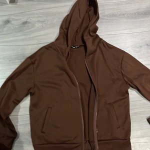 Brun hoodie med dragkedja - Säljer en brun hoodie med dragkedja och huva. Den har långa ärmar och två fickor.