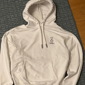 Vit hoodie från Ami - Säljer en stilren vit hoodie från Ami med en liten broderad logga på bröstet. Hoodien har en klassisk design med känguruficka och justerbar dragsko i huvan. Perfekt för en avslappnad look.
