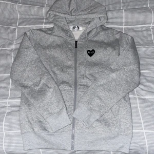 Grå hoodie med hjärtdetalj - Säljer en grå hoodie från Comme des Garçons med dragkedja och huva. Den har ett svart hjärta med ögon på bröstet och ett större på ryggen. Perfekt för en stilren och avslappnad look.