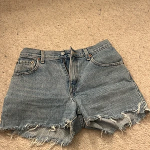 Blå jeansshorts från Levi's - Säljer ett par klassiska blå jeansshorts från Levi's. De har en avslappnad passform med fransiga kanter och är tillverkade i 100% bomull. Perfekta för en avslappnad stil.