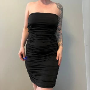 Den lilla svarta  - Snygg svart axelbandslös klänning med tight passform. Klänningen har en elegant rynkad design som ger en smickrande siluett. Perfekt för en kväll ute eller en speciell tillställning. Finnes halterneck knytning som jag vikt in.