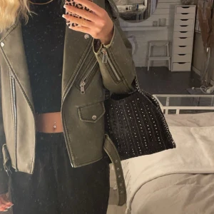 Svart axelväska med nitar - Snygg svart axelväska dekorerad med små nitar. Väskan har en kedja som axelrem och ett stilrent mönster. Perfekt för att ge din outfit en edgy touch.