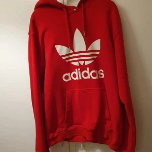 Röd hoodie från Adidas - Säljer en snygg röd hoodie från Adidas med det klassiska trefoil-logotypen i vitt på framsidan. Hoodien har en bekväm passform med långa ärmar och en praktisk känguruficka. Nästan som ny skick. 