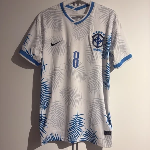 Brasilien nummer 8 (L. Paqueta) - Snygg vit Brasilien fotbollströja från Nike med blå detaljer och palmbladsmönster. Tröjan har nummer 8 och texten 'l. paqueta' på ryggen. Perfekt för fotbollsfans som vill sticka ut med en unik design.