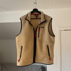 Beige fleeceväst från Patagonia - Beige fleeceväst från Patagonia i storlek L. Västen har en dragkedja framtill och praktiska fickor med dragkedjor. Den är perfekt för kyligare dagar och har en stilren design med röda och mörkblå detaljer.