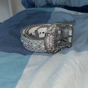 Glittrigt bälte med strass. Lite sönder på ett ställe men det är bara på kanten. - Säljer ett glittrigt bälte i silver med massor av strassdetaljer. Bältet har en stor, dekorerad spänne och är perfekt för att ge lite extra bling till din outfit. Det är kantat med små pärlor längs kanterna för en extra touch av glamour.
