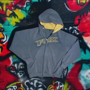 Grå hoodie från Fox - Säljer en grå hoodie från Fox med gul insida av huvan och ett stort tryck på framsidan. Tröjan har dragkedja och långa ärmar, perfekt för en avslappnad stil. 🦊