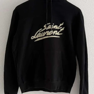 Svart hoodie från Saint Laurent - Snygg svart hoodie från Saint Laurent med vit logga på bröstet. Tröjan har en klassisk design med huva och dragsko samt en stor magficka framtill. Perfekt för en stilren och avslappnad look.