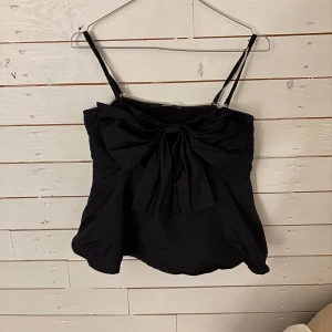 Svart topp med rosett - Snygg svart axelbandslös topp från BikBok med en stor rosett framtill. Perfekt för en kväll ute eller en speciell tillställning. Toppen har justerbara axelband för bästa passform.